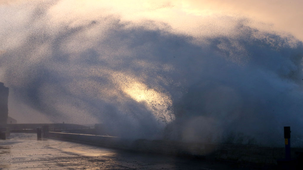 A wave breaks over the seafront at dawn in West Bay, Dorset (G)
